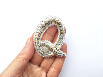 A hand holding a decorative letter 'Q' made of beads, pearls, and other embellishments.