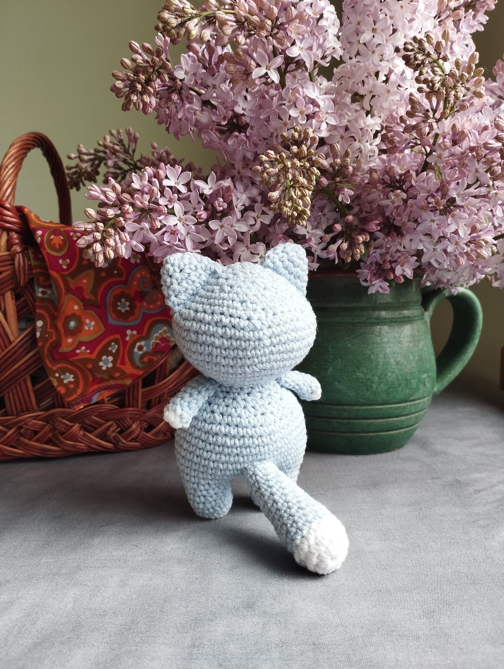 a crocheted teddy bear sitting next to a basket of lilacs