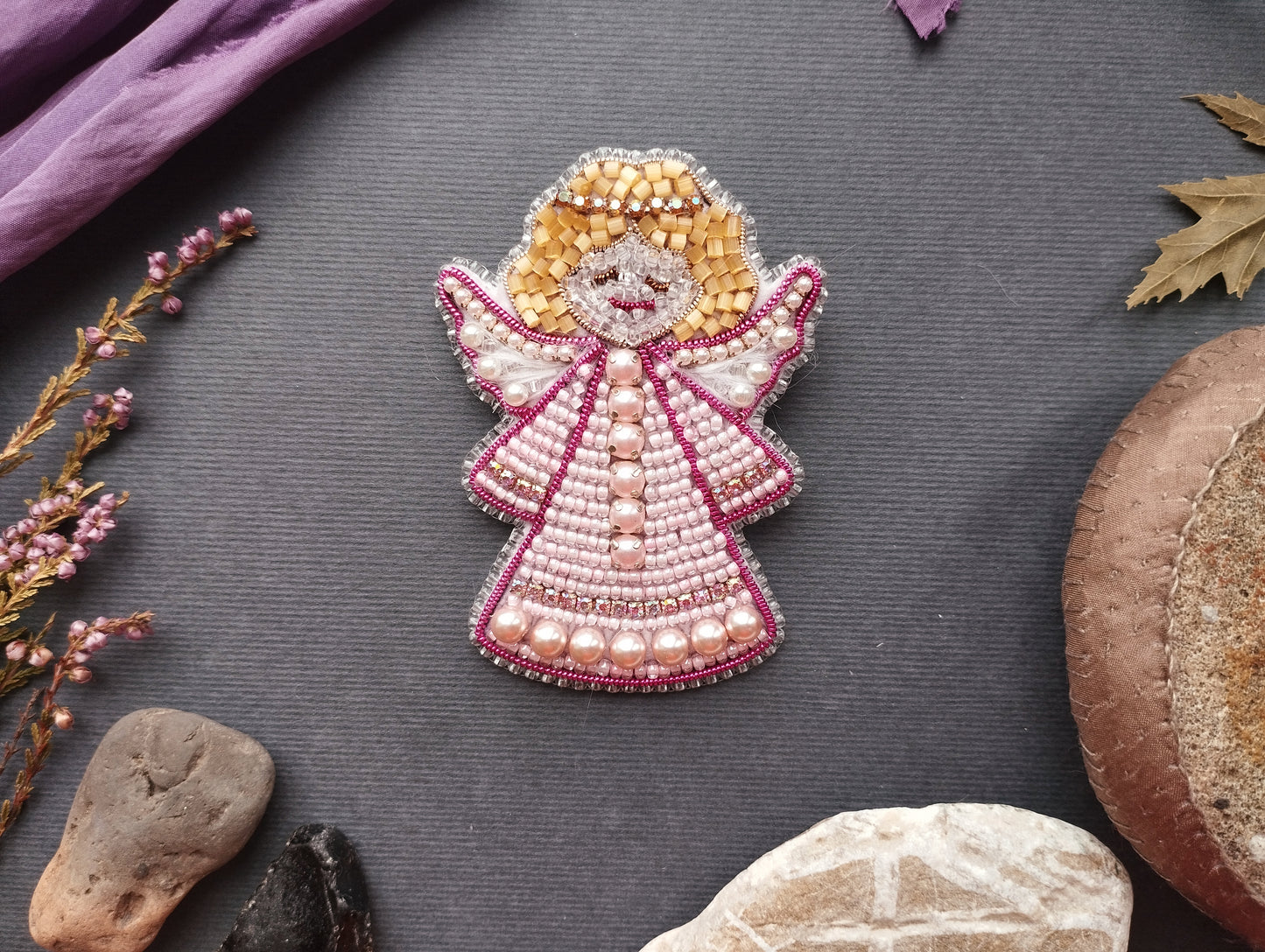 a decorative piece of art, possibly a beaded or embroidered design, placed on a grey surface surrounded by various natural elements such as leaves, flowers, and rocks.