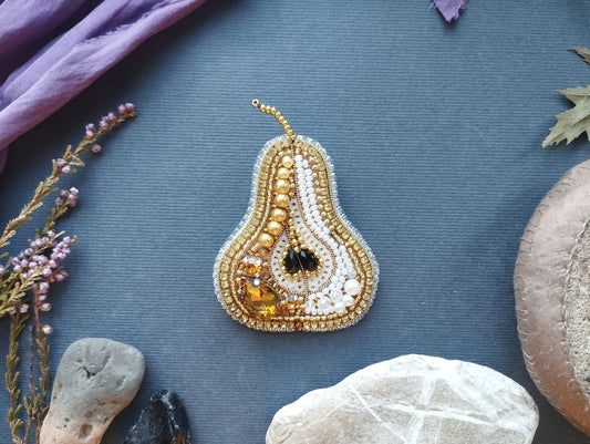a decorative piece of jewelry, specifically a pear-shaped brooch, which is adorned with various materials such as beads, stones, and leaves. The pear-shaped brooch is placed on a blue surface, surrounded by other natural elements like rocks and leaves.