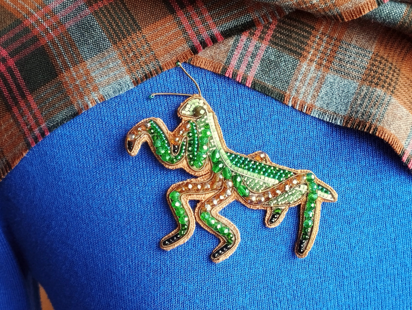 A close-up image of a decorative green and gold embroidered insect, resembling a grasshopper or cricket, on a blue fabric surface.