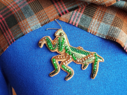 A close-up image of a decorative green and gold embroidered insect, resembling a grasshopper or cricket, on a blue fabric surface.