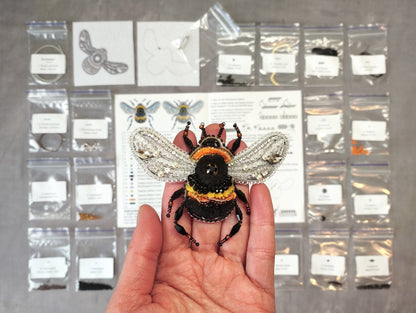A hand is holding a large, intricately designed beaded bee pin up on its fingers, with a variety of small bags and packages containing different items, such as beads and other craft supplies, visible in the background.