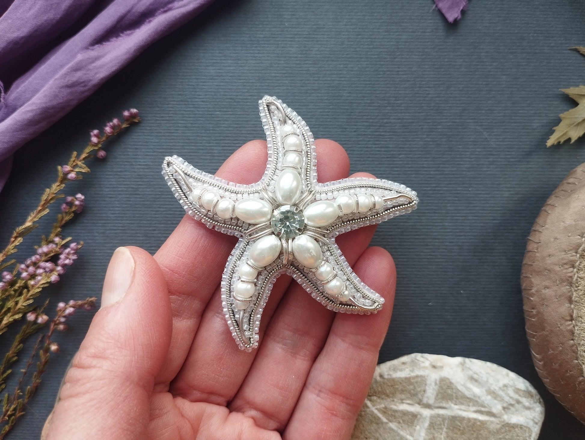 A hand holding a decorative starfish with pearls and a gemstone on a dark background.