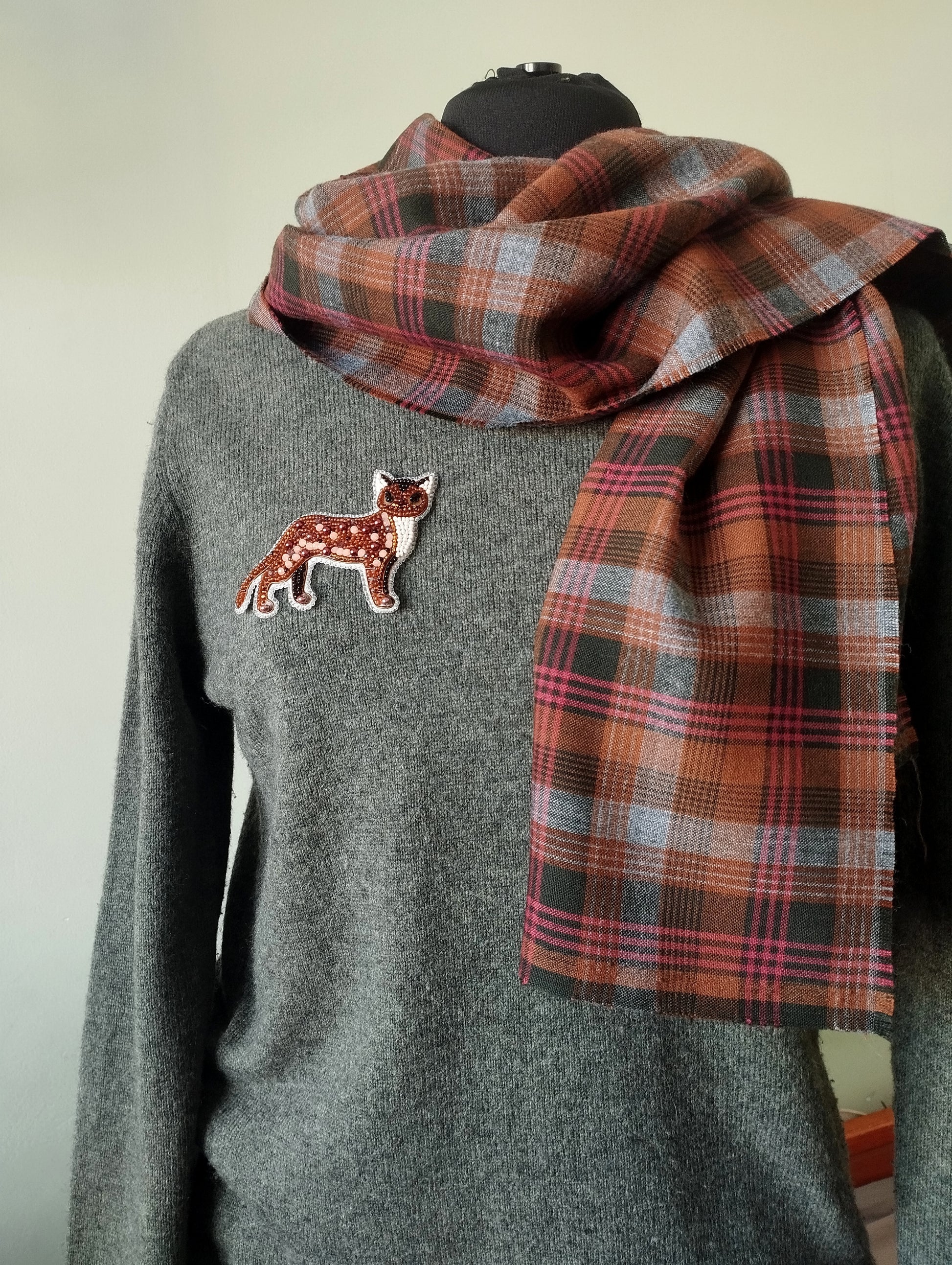 A gray sweater with a patch of a cat embroidered on it, and a plaid scarf draped over the sweater.