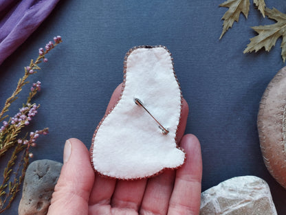 A hand holding a small, white, felt-like object with a pin or pinwheel attached to it, surrounded by various natural elements such as leaves, flowers, and rocks.