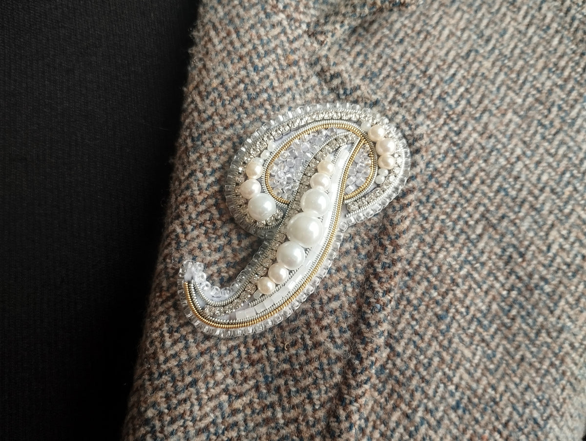 A close-up image of a decorative monogram brooch or pin, featuring a combination of pearls, beads, and metallic elements, attached to a fabric surface.