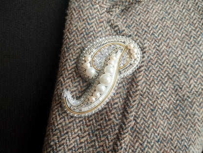 A close-up image of a decorative monogram brooch or pin, featuring a combination of pearls, beads, and metallic elements, attached to a fabric surface.