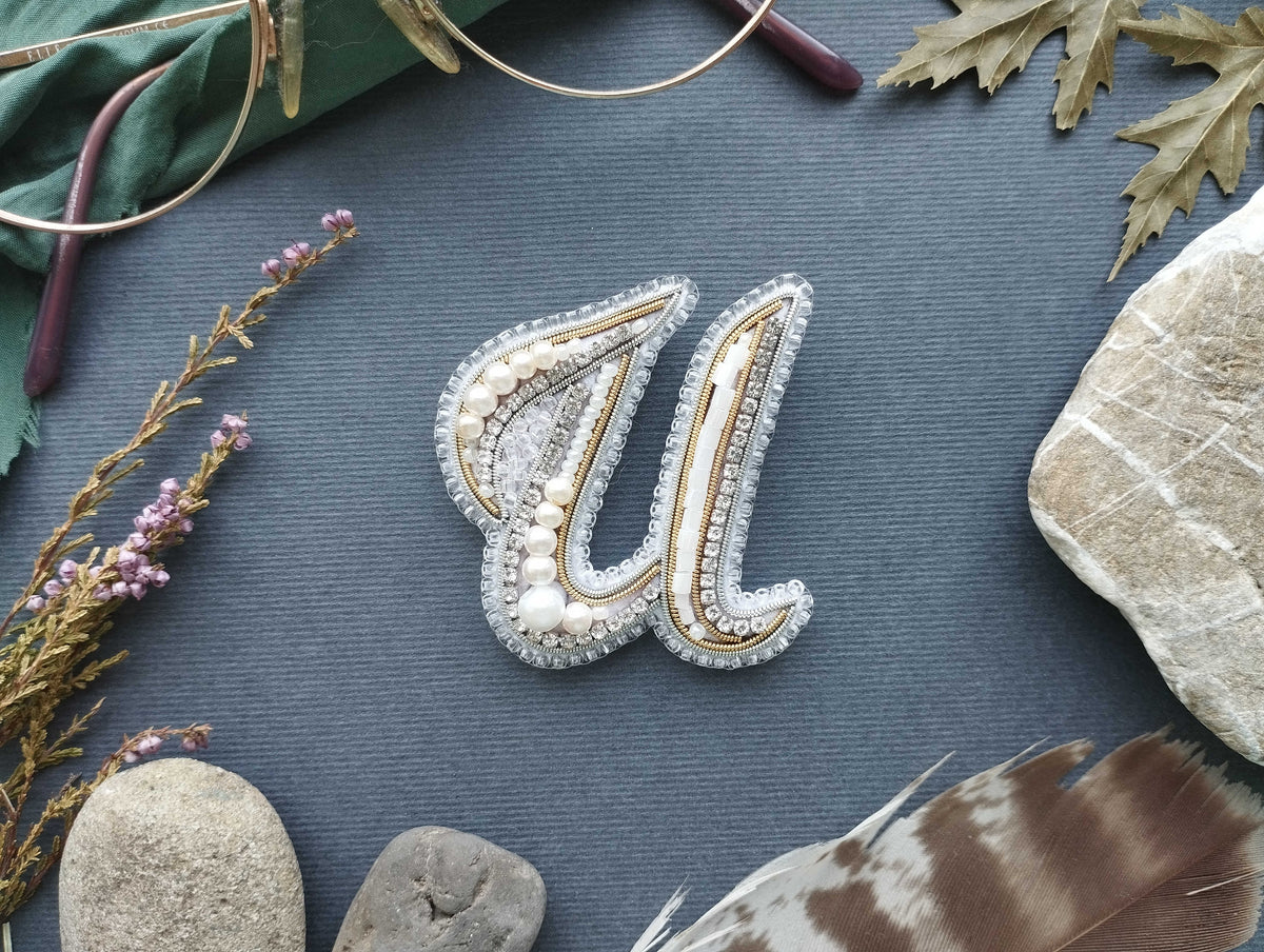 a close-up of a decorative letter 'U' made of beads, pearls, and other embellishments, surrounded by various natural elements such as feathers, rocks, and flowers.
