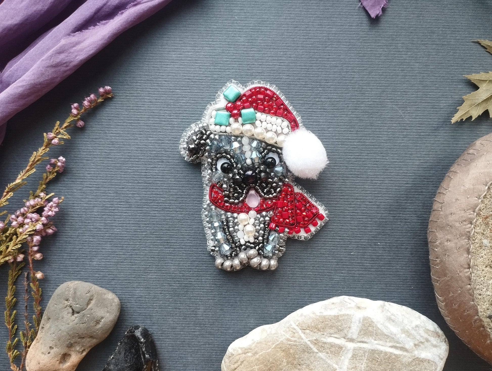a small, intricately designed pin or brooch in the shape of a dog, adorned with red and silver beads and a red and white pom pom. The pin is placed on a gray surface, surrounded by various objects such as rocks, leaves, and flowers.