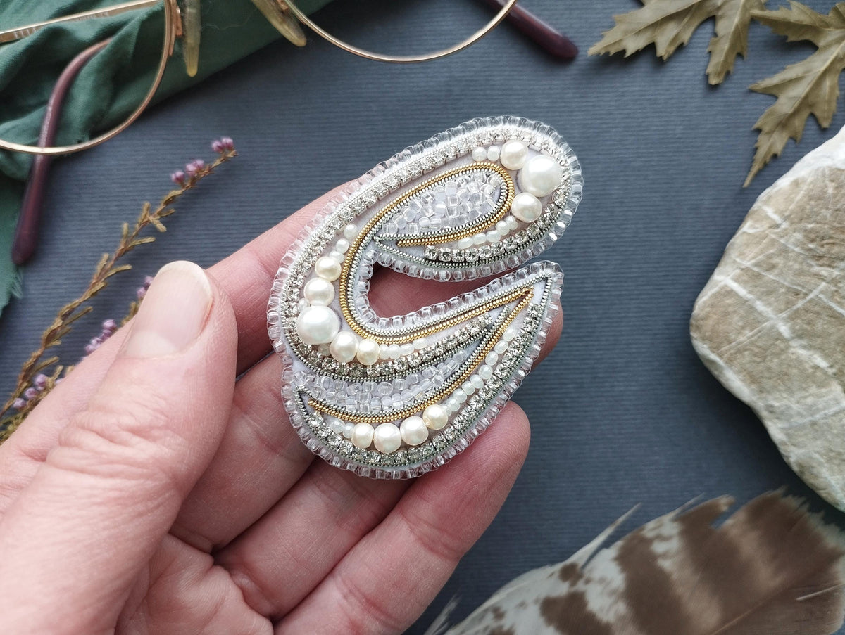 A hand holding a decorative piece of jewelry, which appears to be a brooch or pin, against a backdrop of various natural elements such as leaves, feathers, and rocks.