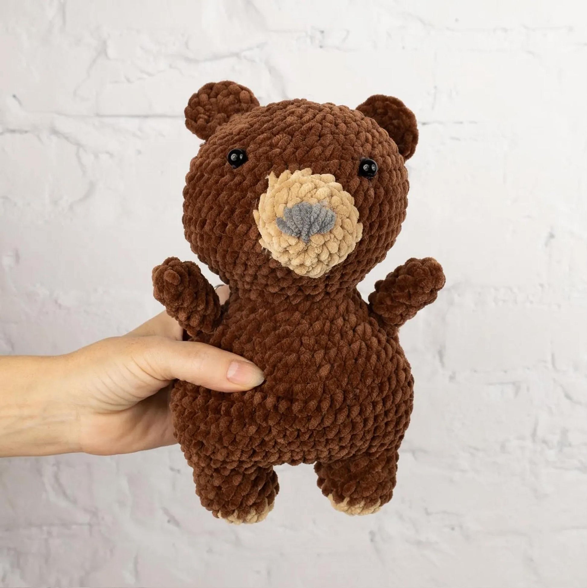 A hand is holding a brown, crocheted stuffed bear toy against a white brick wall background.