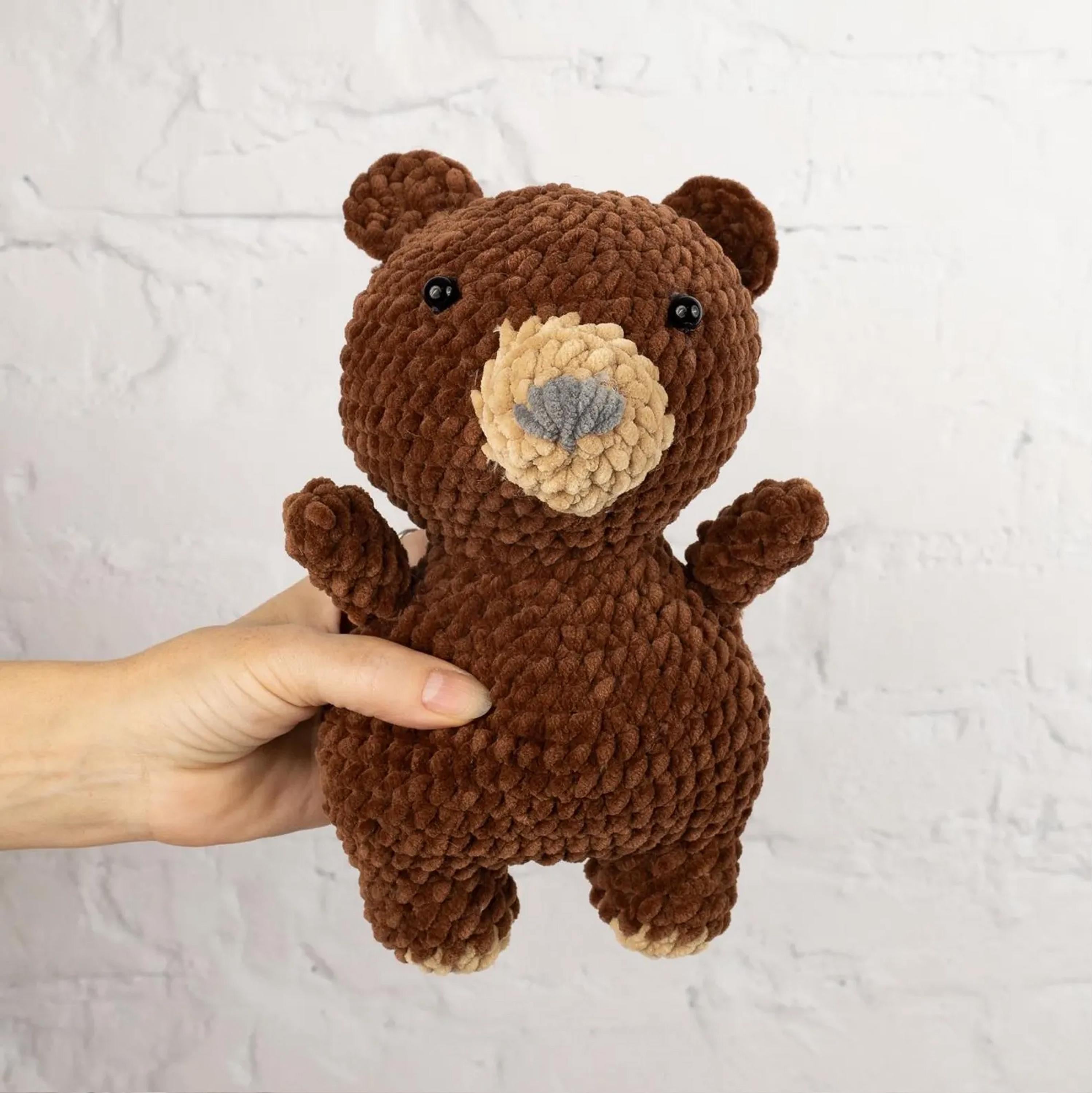 A hand is holding a brown, crocheted stuffed bear toy against a white brick wall background.