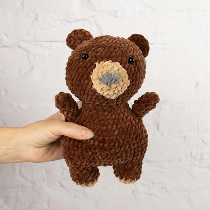 A hand is holding a brown, crocheted stuffed bear toy against a white brick wall background.