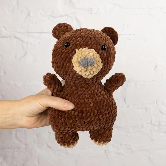A hand is holding a brown, crocheted stuffed bear toy against a white brick wall background.