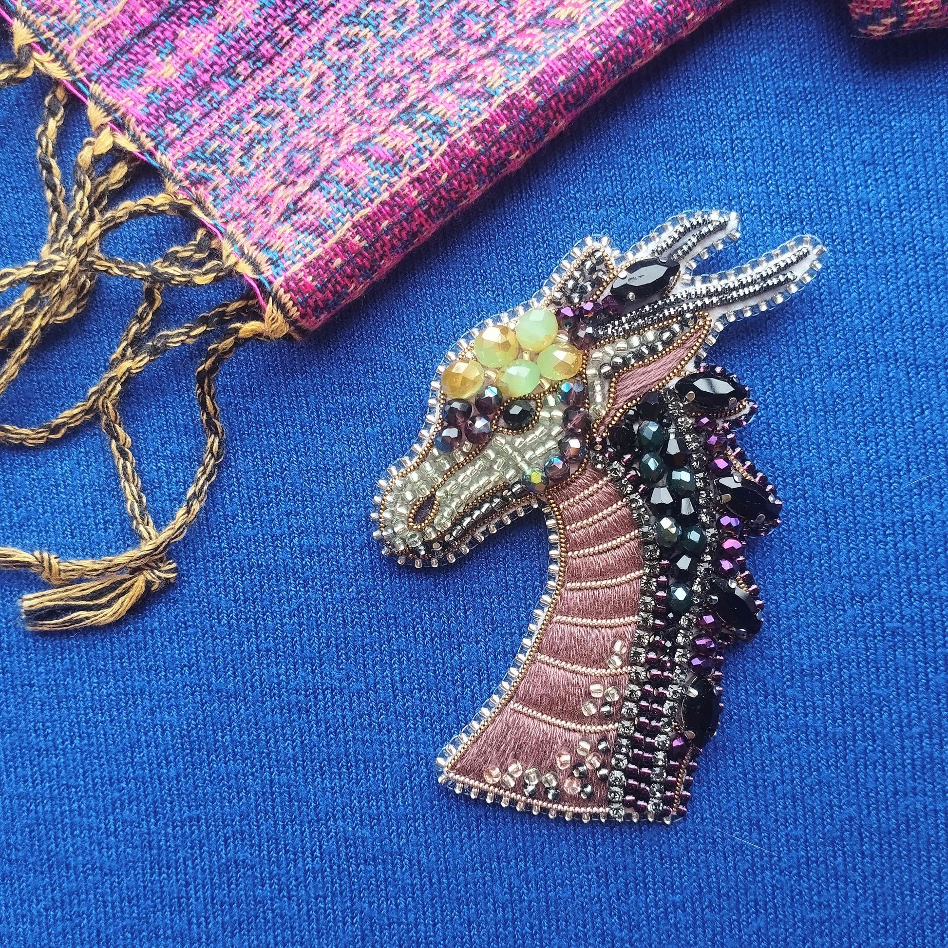 a close-up view of a decorative dragon head pin, adorned with colorful beads and sequins, resting on a blue fabric surface.