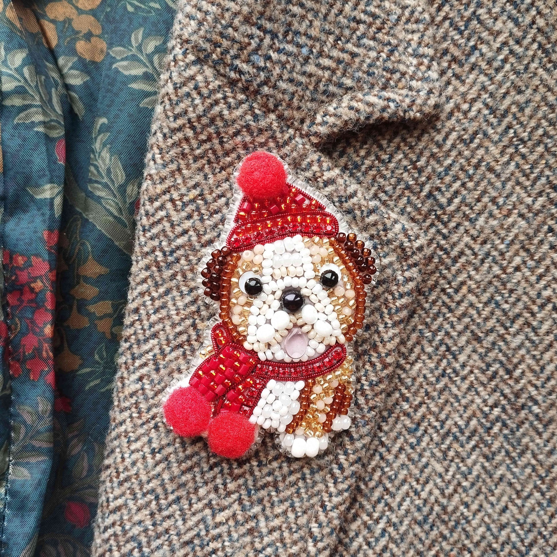 A close-up of a small, intricately beaded dog-shaped brooch or pin, wearing a red hat and scarf, attached to a tweed jacket.