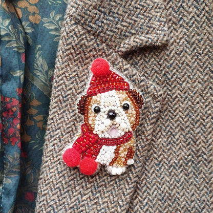 A close-up of a small, intricately beaded dog-shaped brooch or pin, wearing a red hat and scarf, attached to a tweed jacket.