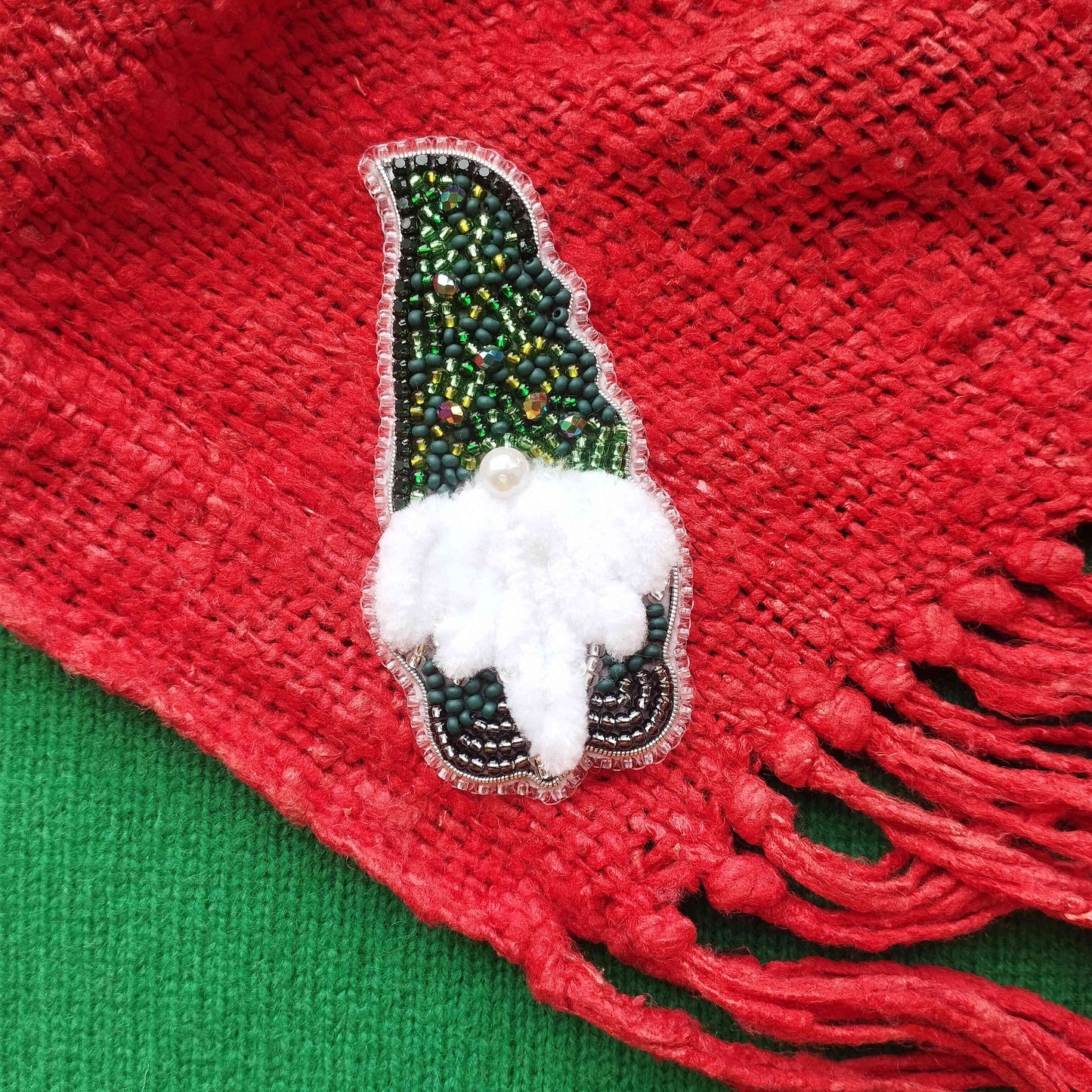 A close-up image of a red fabric with a decorative green and white beaded ornament, resembling a small elf or gnome-like figure, placed on top of it.