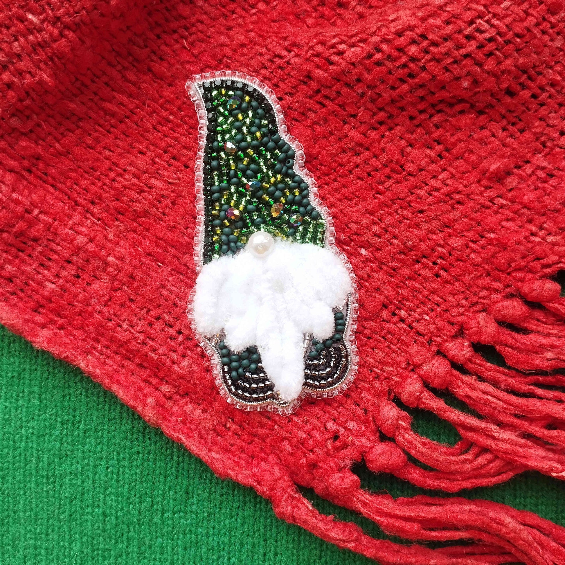 A close-up image of a red fabric with a decorative green and white beaded ornament, resembling a small elf or gnome-like figure, placed on top of it.