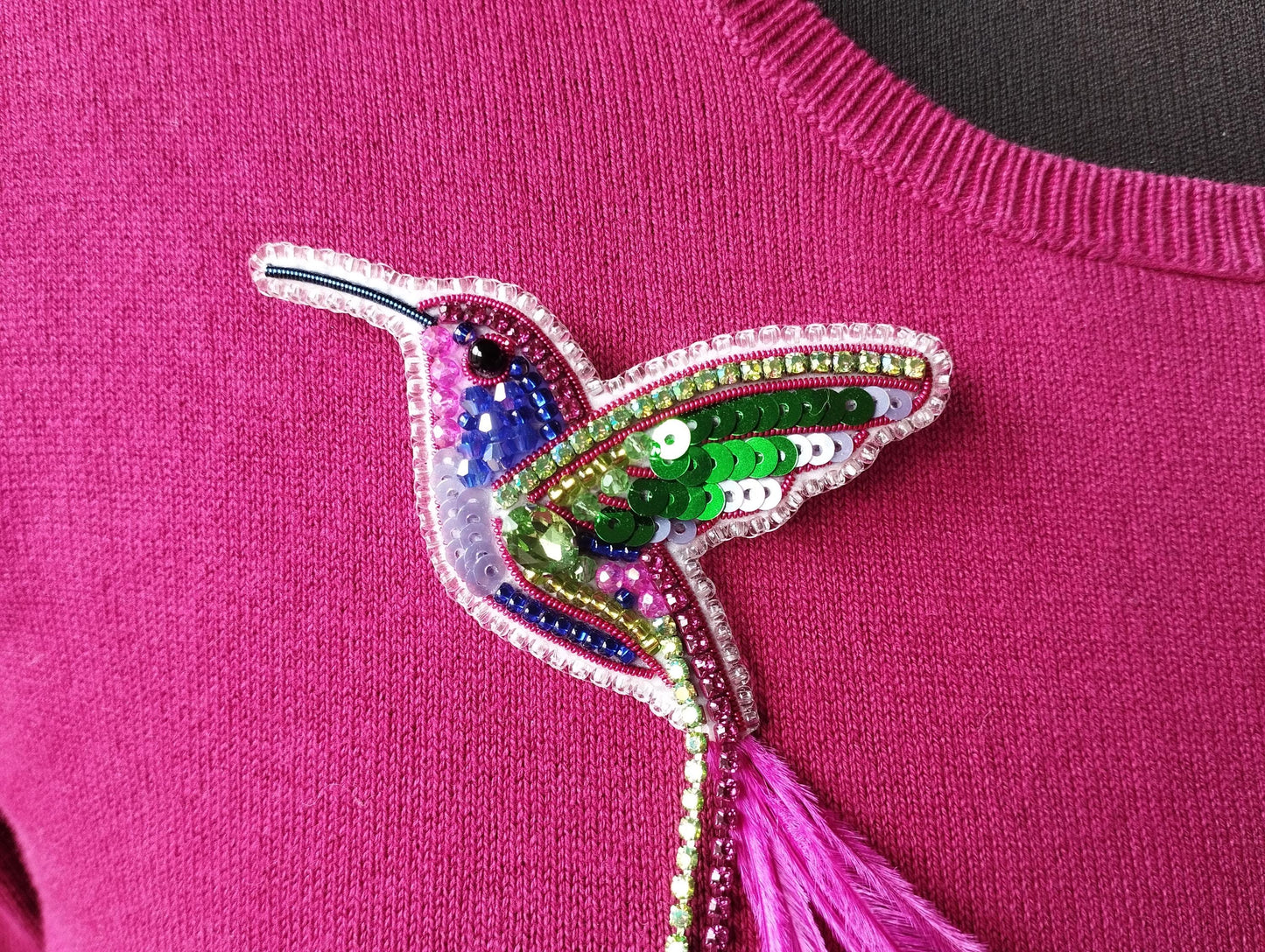 A close-up image of a pink sweater with a colorful, sequined hummingbird brooch or pin attached to it.