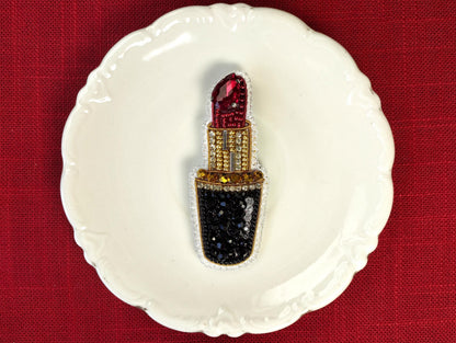 A white plate with a decorative design, featuring a red and gold brooch-like object with a black and gold base, placed on a red surface.