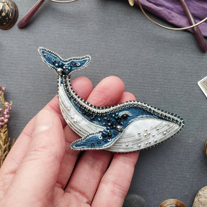 A hand holding a decorative blue and white embroidered piece resembling a whale, with various other objects in the background.
