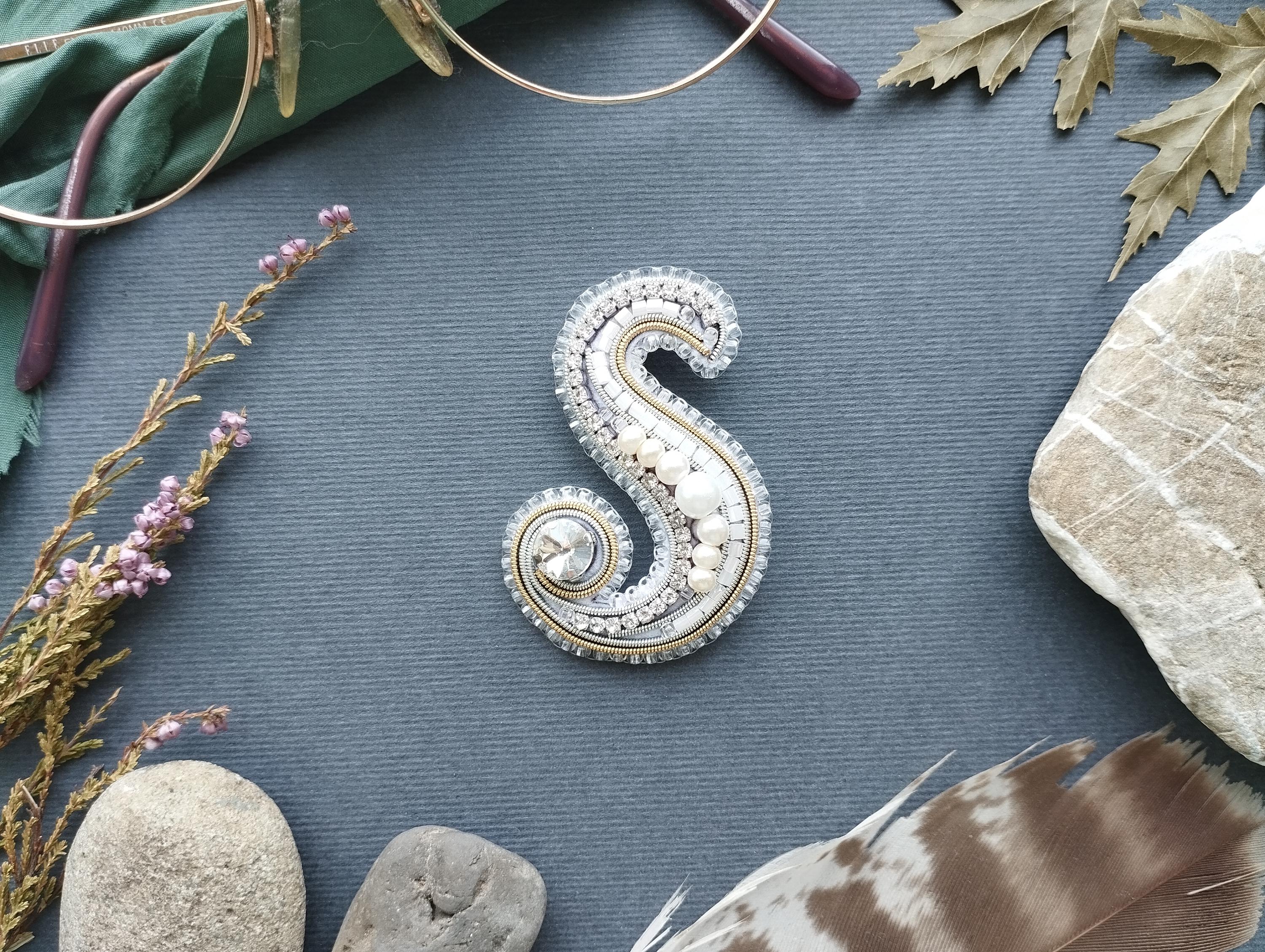 a decorative letter 'S' made of various materials, including jewels and feathers, arranged on a blue background.