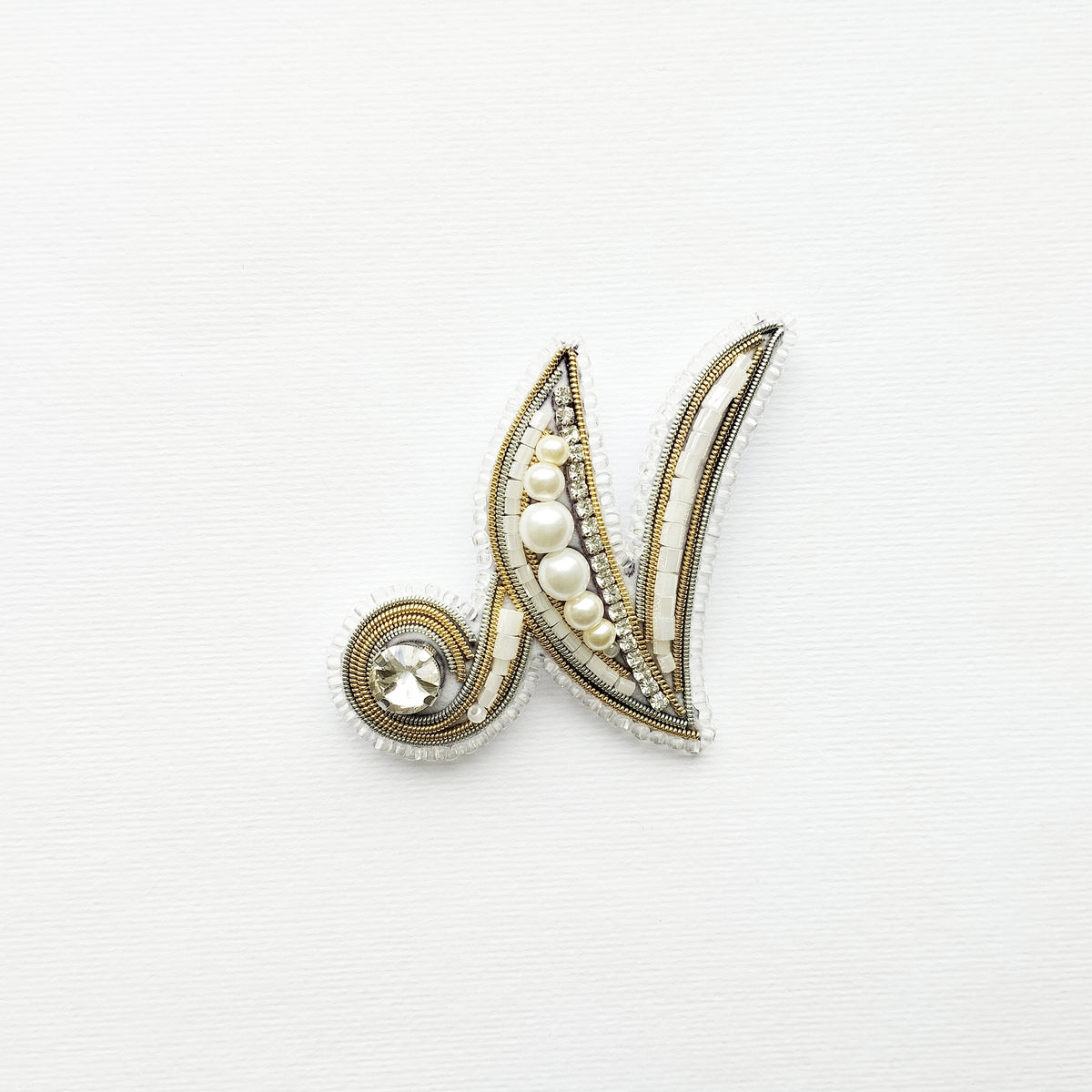 A decorative letter 'M' made of gold, pearls, and crystals against a white background.