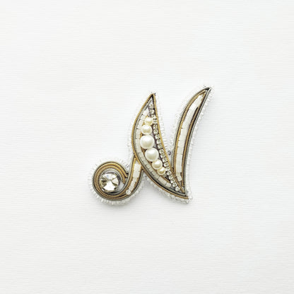 A decorative letter 'M' made of gold, pearls, and crystals against a white background.