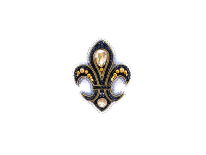 A decorative black and gold emblem featuring a fleur-de-lis design with a central gemstone surrounded by smaller gemstones and pearls.