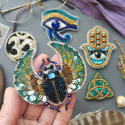 a hand holding a decorative piece of jewelry, which appears to be a beaded or embroidered design. The hand is holding a large, intricately designed piece that resembles a butterfly or a dragonfly. The background includes various other pieces of jewelry, such as a hamsa charm, a hand of faith charm, and a trident charm, all of which are also adorned with colorful beads and sequins.