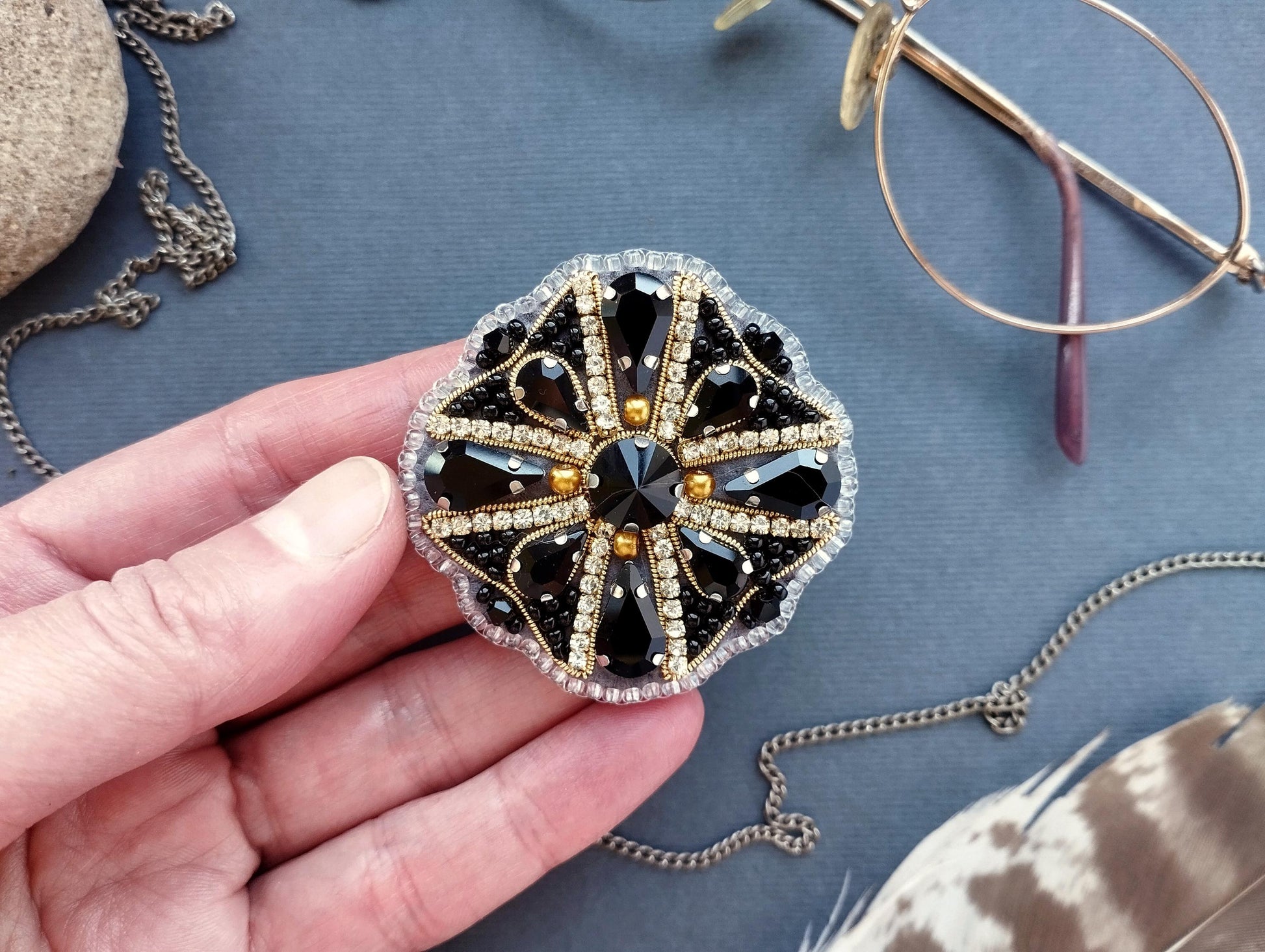 A hand holding a decorative piece of jewelry, which appears to be a brooch or pendant, against a blue background with various accessories such as glasses and feathers.
