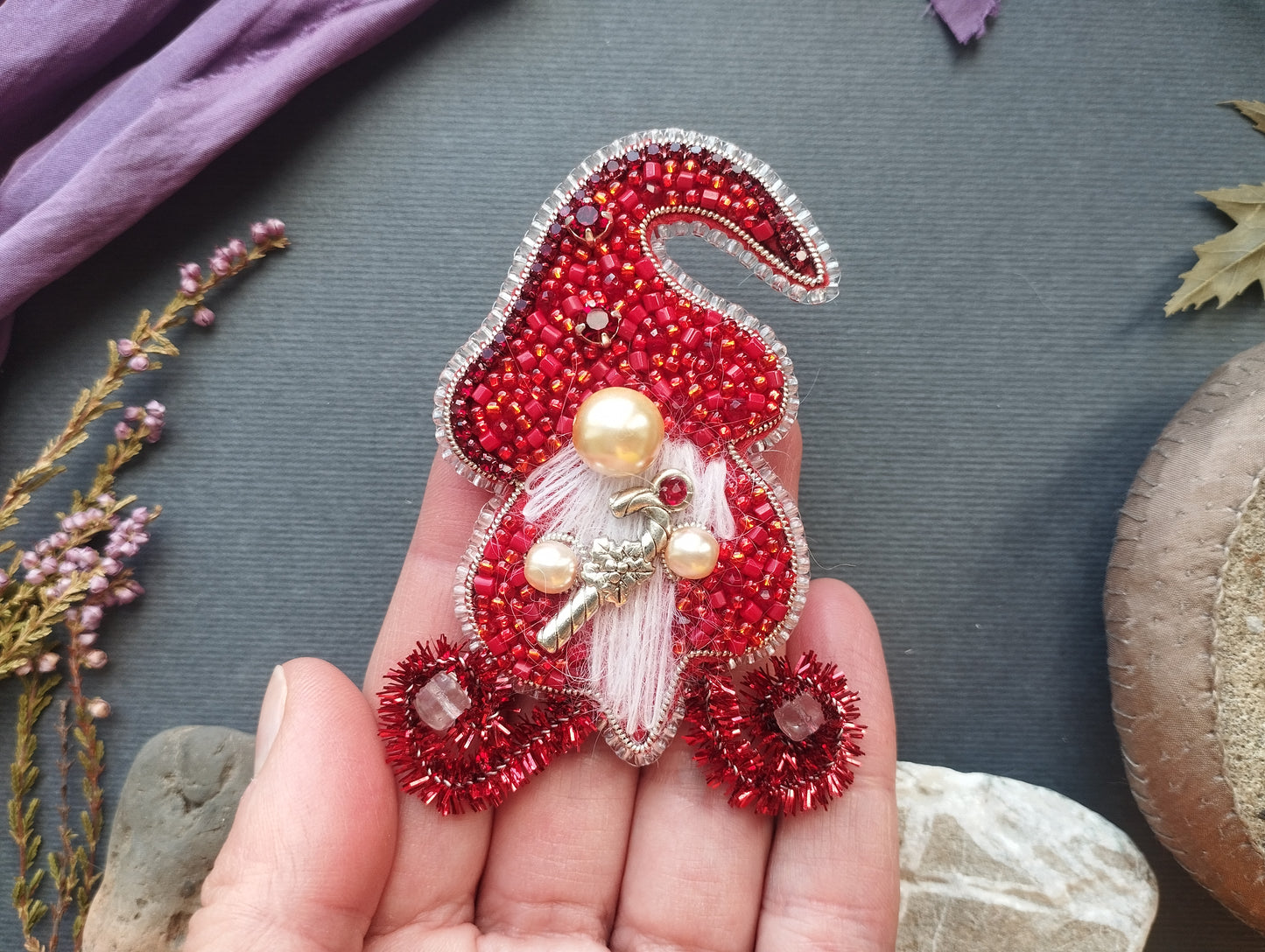 A hand holding a red and silver ornament with a gnome-like design, adorned with red and gold beads, pearls, and a key.