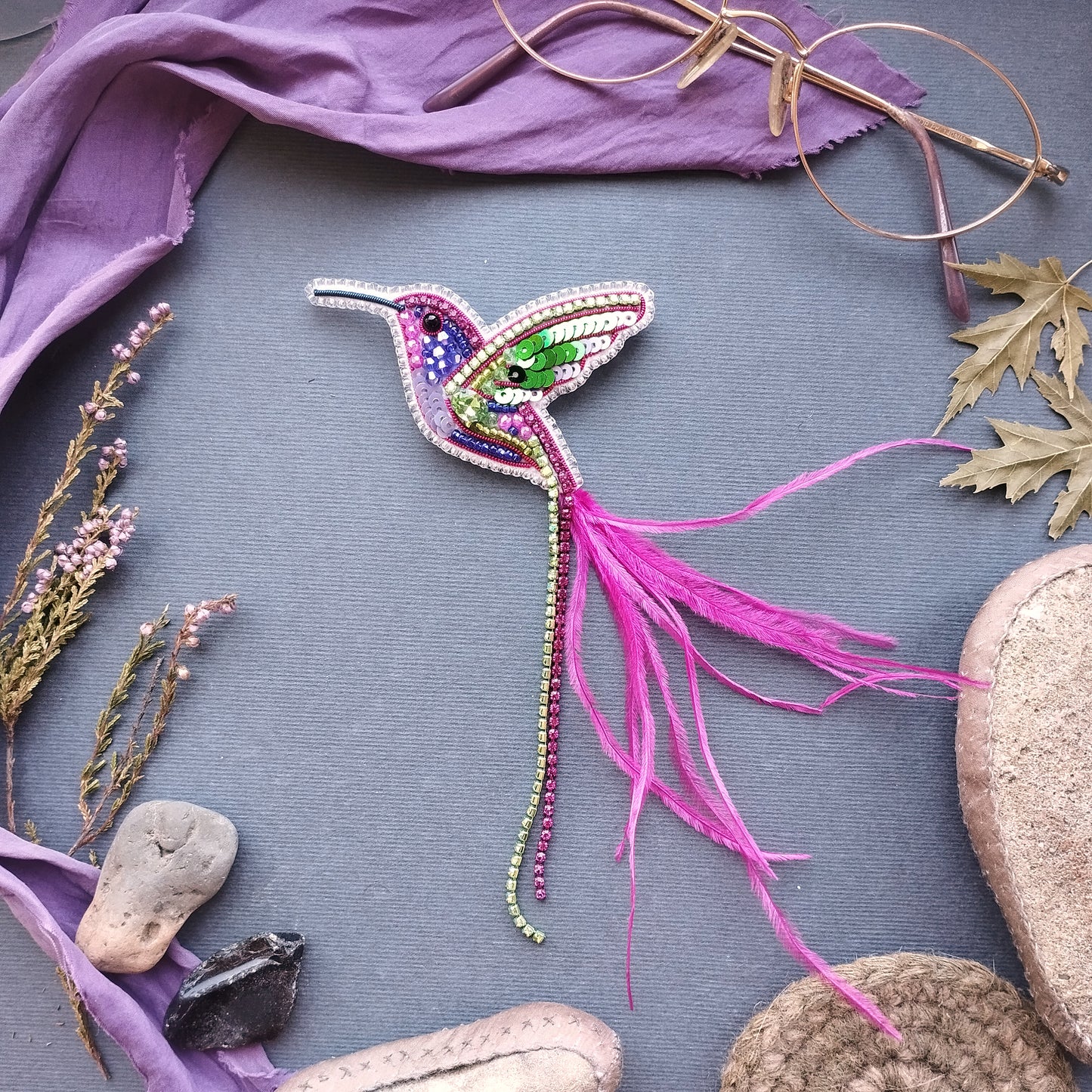a colorful, hand-made hummingbird brooch with a long pink feather, surrounded by various natural elements such as leaves, flowers, and rocks.