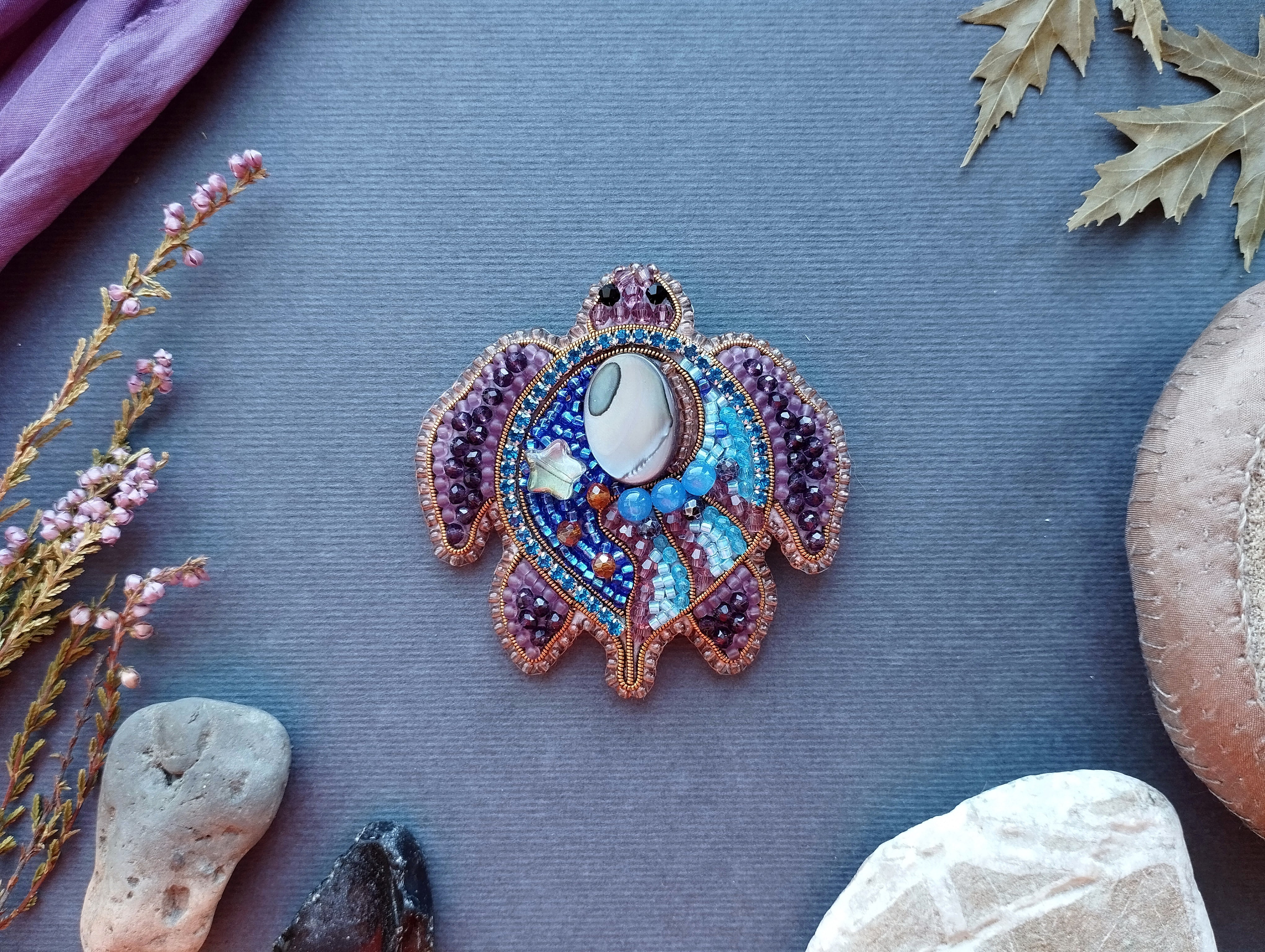 a decorative turtle brooch with a blue and purple color scheme, adorned with various beads and stones, placed on a blue surface.