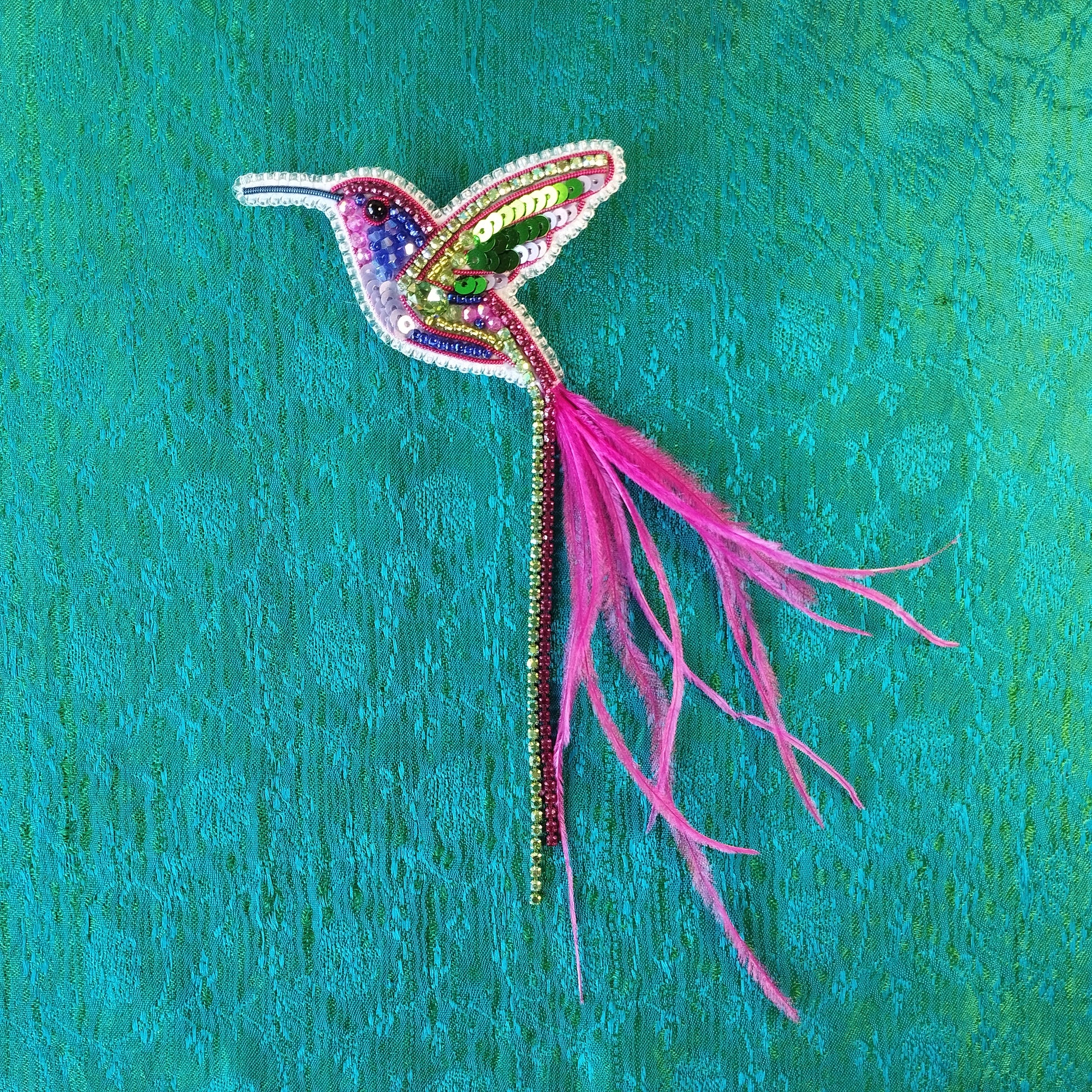 A colorful, intricately designed hummingbird brooch made of beads and feathers is displayed against a vibrant green background.