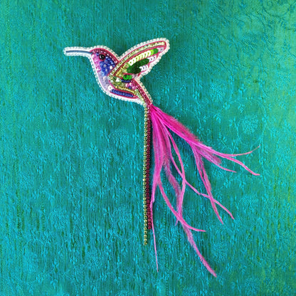 A colorful, intricately designed hummingbird brooch made of beads and feathers is displayed against a vibrant green background.