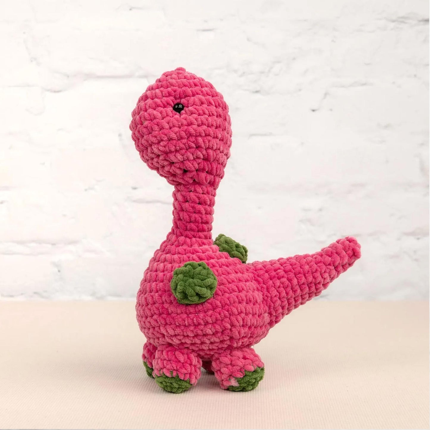 A pink crocheted dinosaur toy is sitting on a white surface against a white brick wall background.