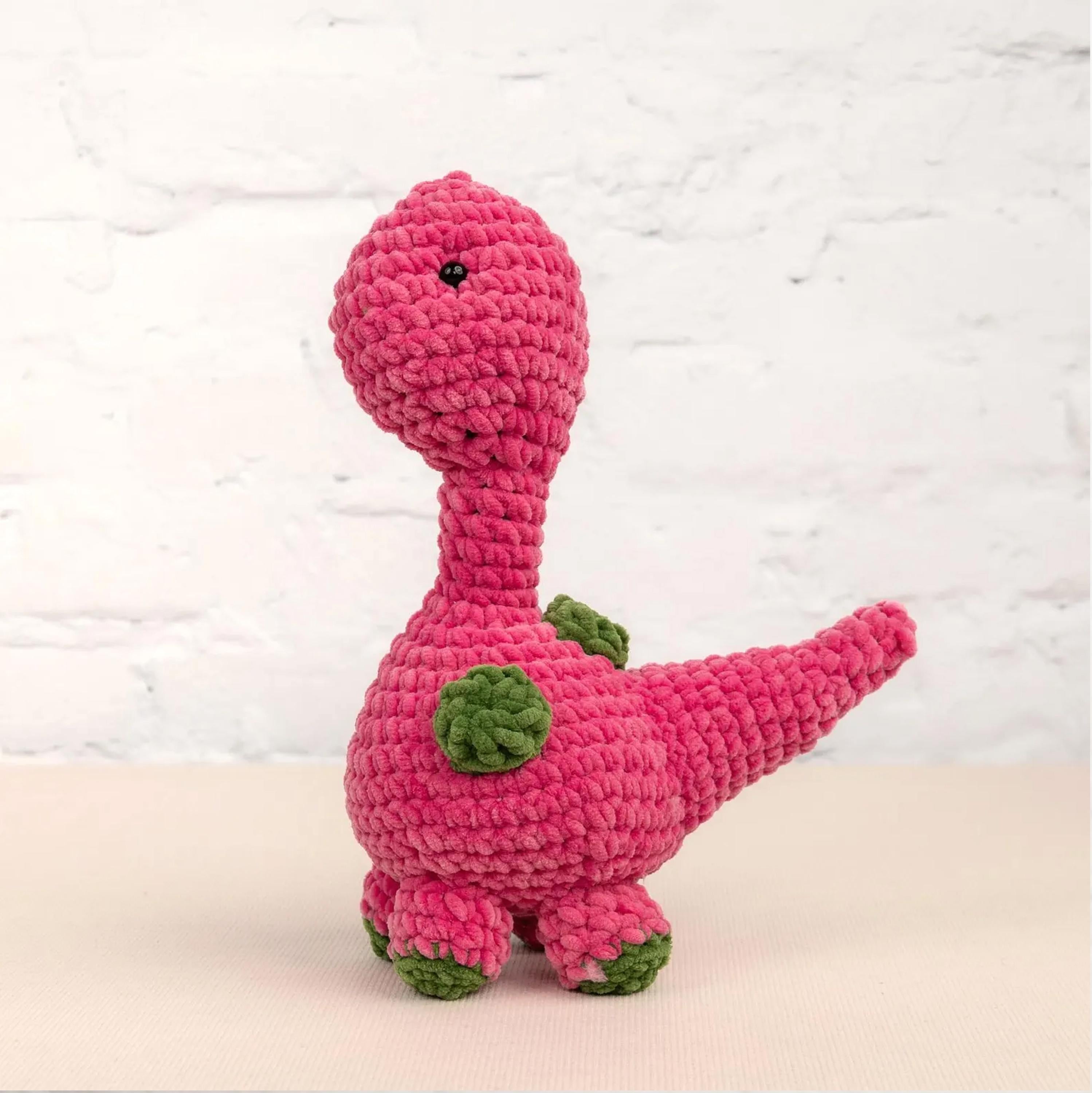 A pink crocheted dinosaur toy is sitting on a white surface against a white brick wall background.