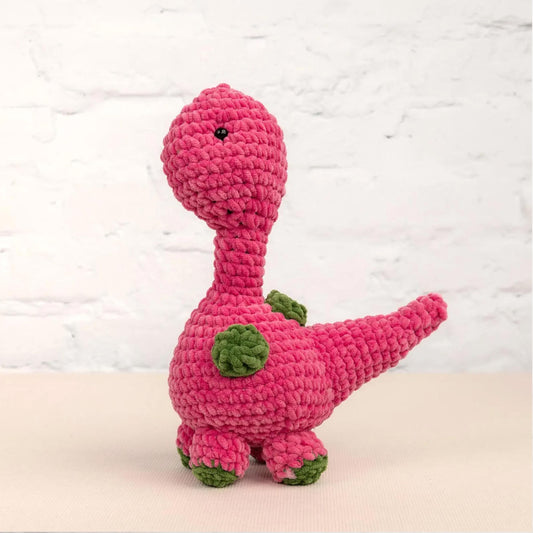 A pink crocheted dinosaur toy is sitting on a white surface against a white brick wall background.