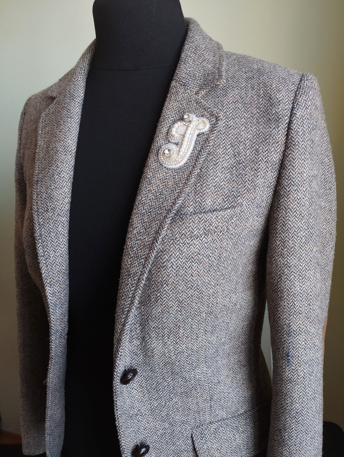 A gray tweed blazer with a white embroidered logo on the left side of the chest.