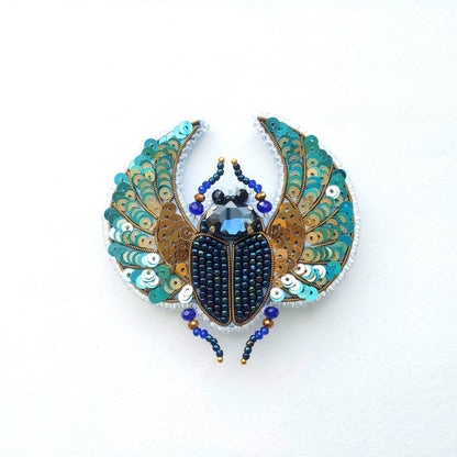 A decorative piece featuring a detailed embroidered design of a colorful insect, possibly a beetle or a fly, with intricate patterns and embellishments.