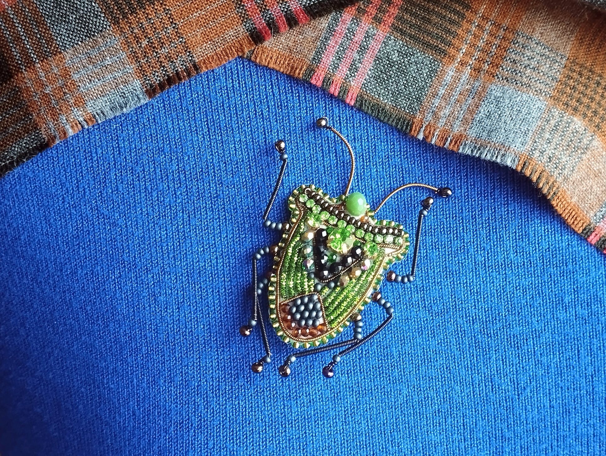 A colorful, intricately designed insect brooch is displayed against a blue fabric background, with a plaid fabric visible in the top right corner.