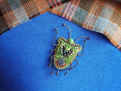A colorful, intricately designed insect brooch is displayed against a blue fabric background, with a plaid fabric visible in the top right corner.