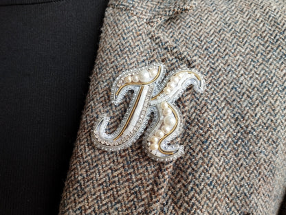 A close-up image of a decorative monogrammed letter "J" on a tweed fabric, with pearls and rhinestones embellishing it.