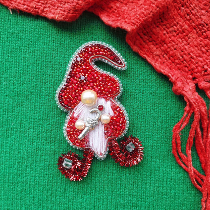 A red and silver ornament resembling a gnome or elf is displayed on a green background, with a red scarf visible in the top right corner.
