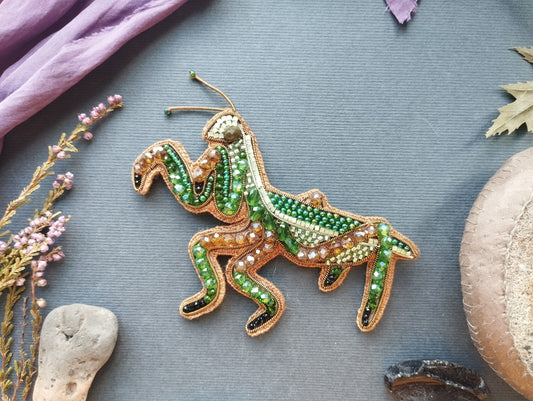 a decorative piece of embroidered grasshopper-shaped jewelry, adorned with green and gold beads, placed on a gray surface alongside various natural elements such as leaves and flowers.