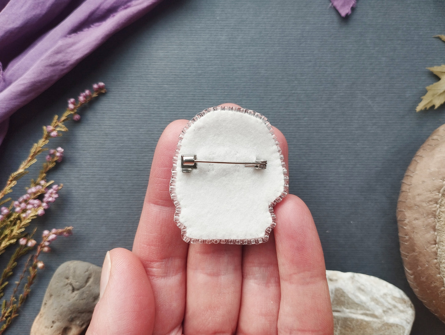 A hand holding a white fabric pin with a silver pinback, surrounded by various objects such as flowers, rocks, and a purple fabric.