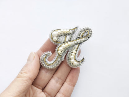 A hand holding a decorative letter 'H' made of beads and pearls.